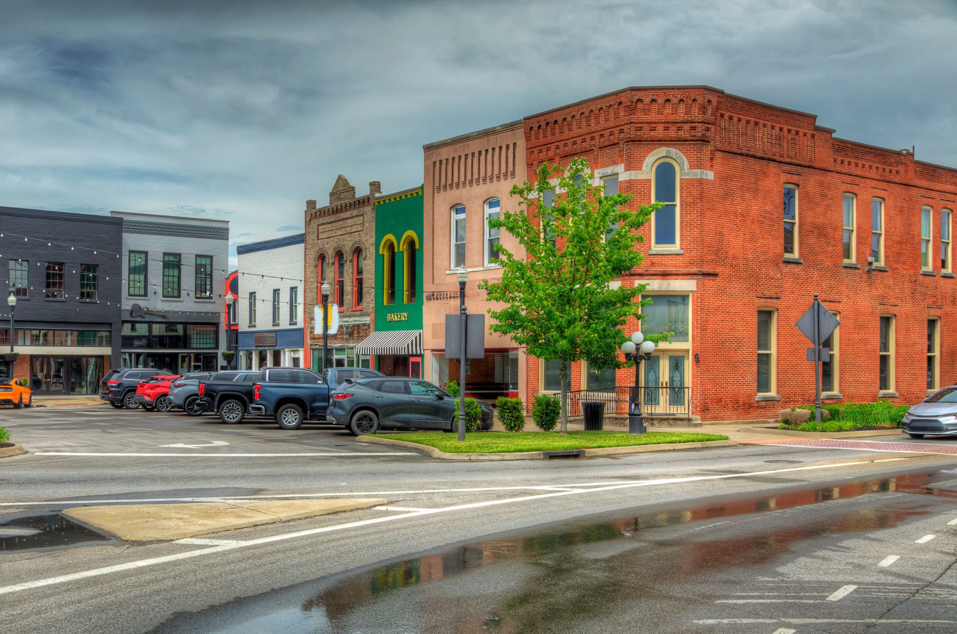 Buildings in various colors line a street. Cars parked along side. Wet pavement reflects the buildings and overcast sky.