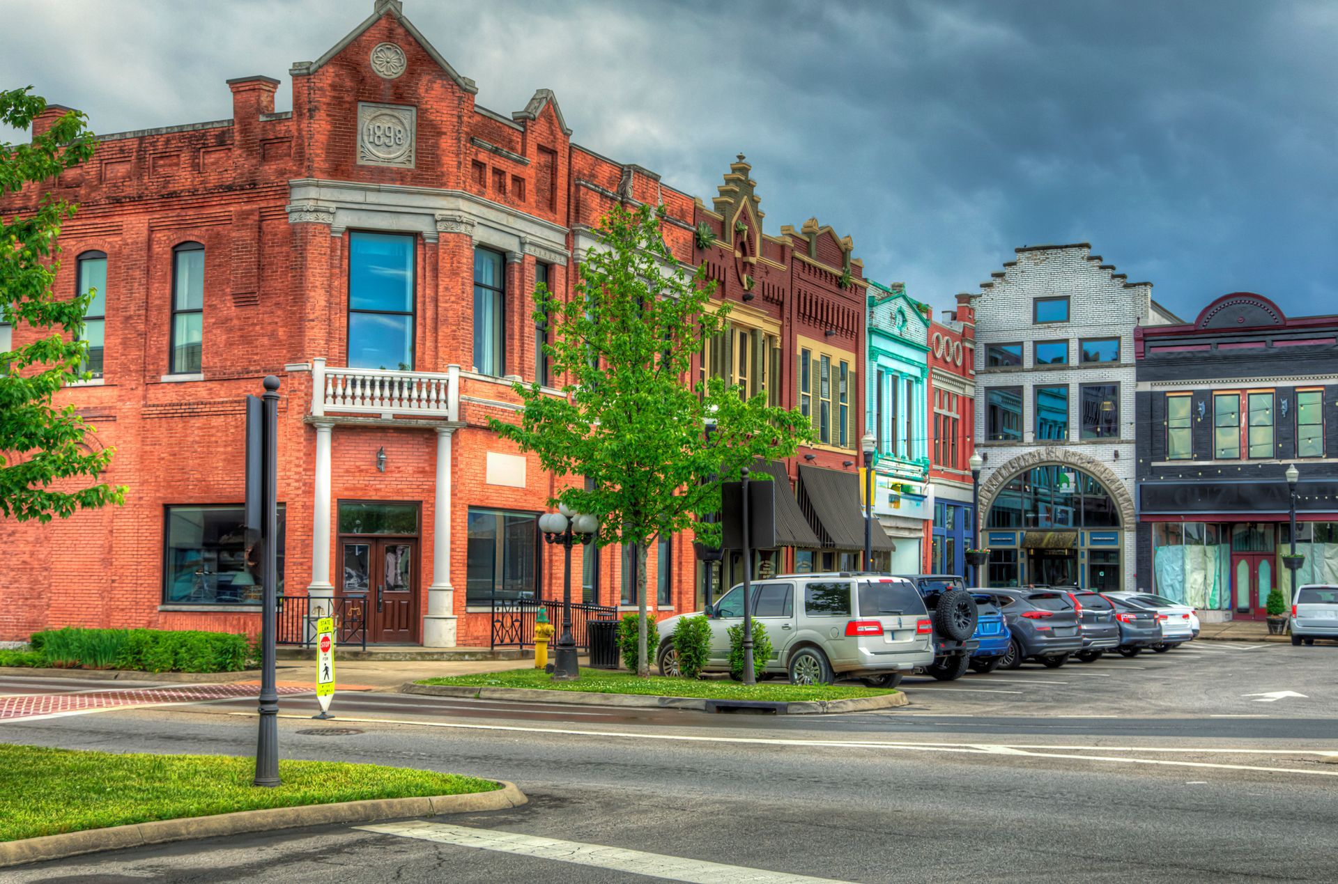 Row of brick and colorful buildings along a street with parked cars and overcast sky.