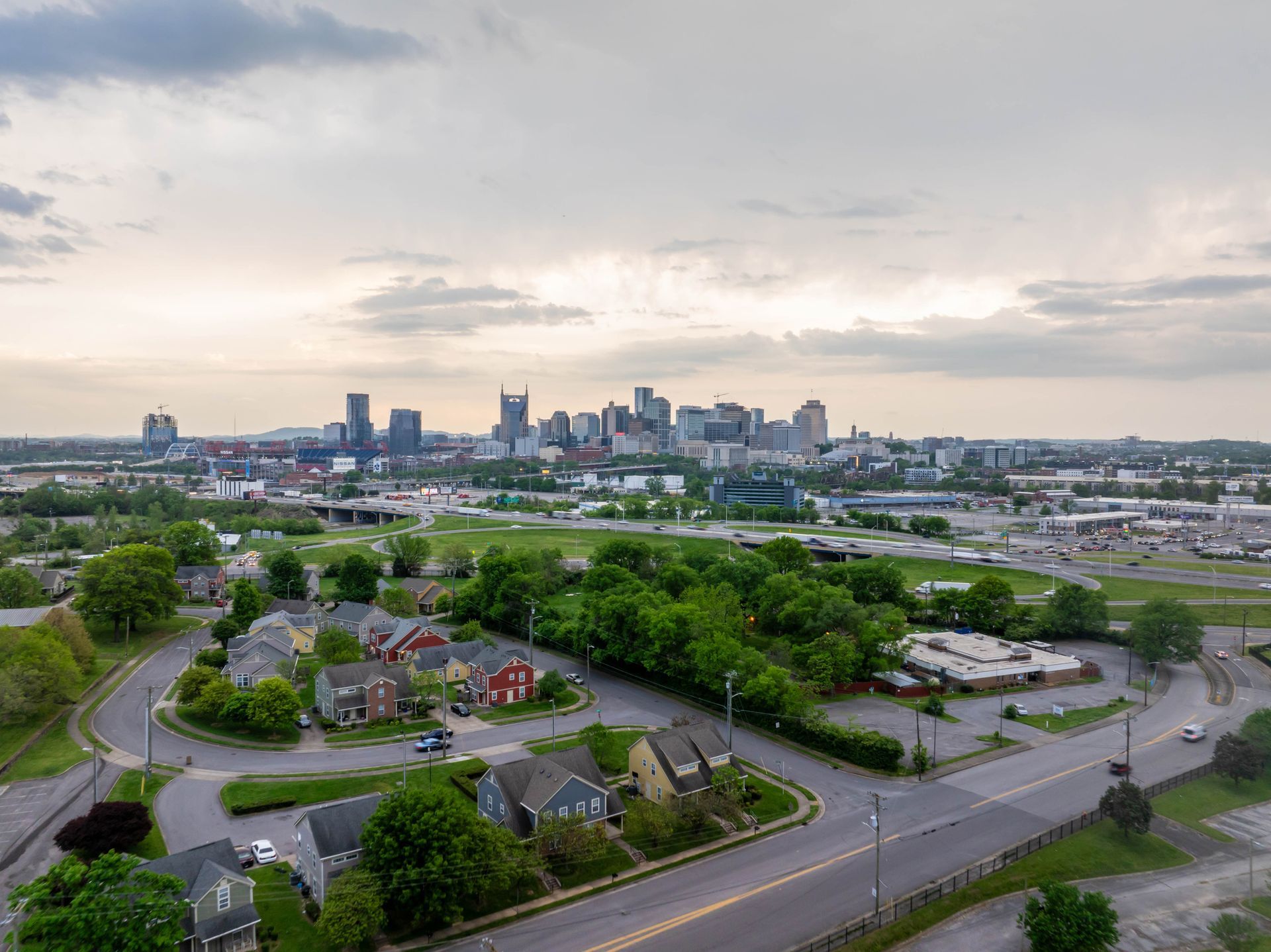 City skyline at dusk, viewed from above a residential area. Buildings and road with green trees under a cloudy sky.