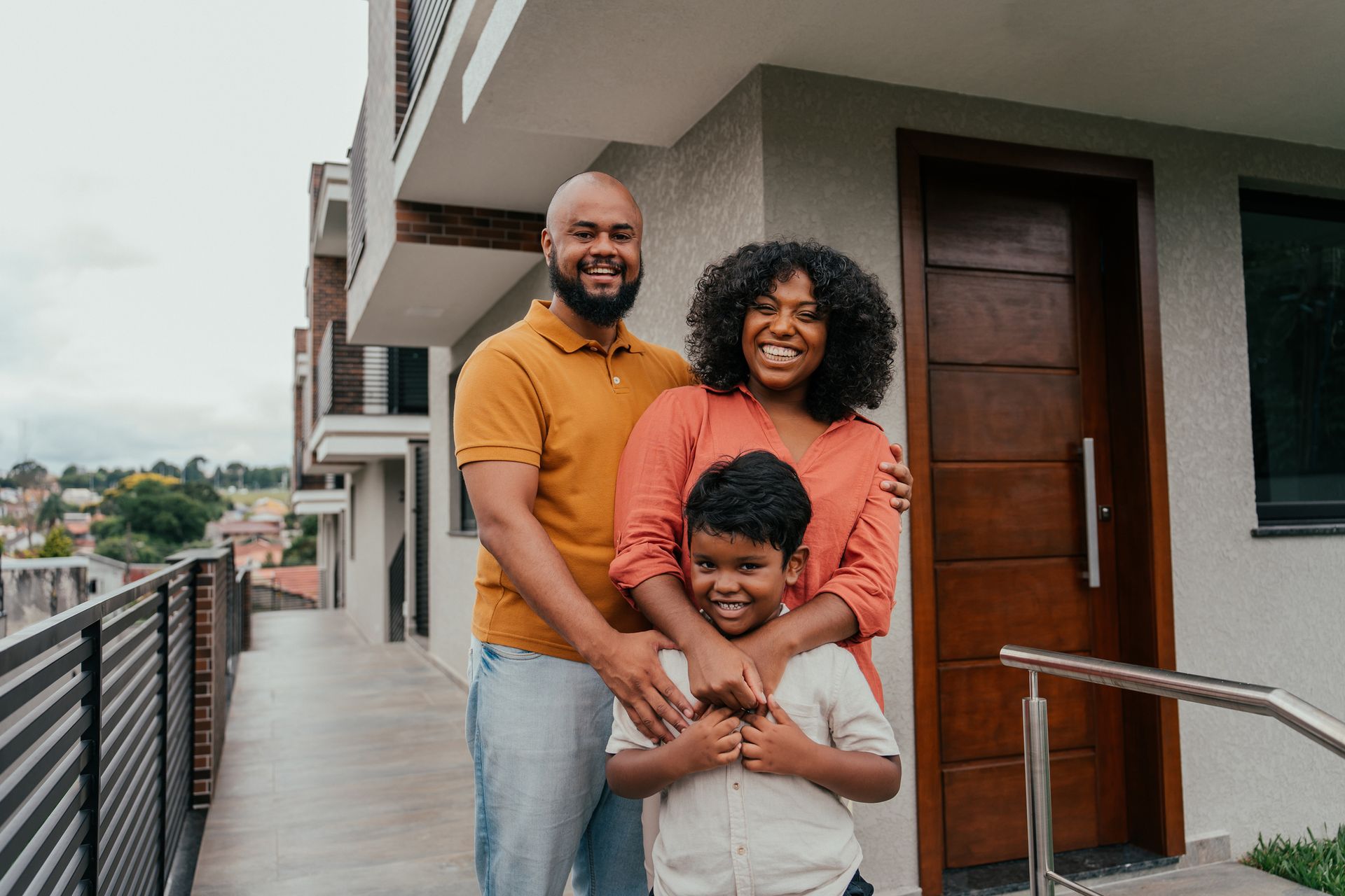 Family of three smiling in front of a modern home; man, woman, child.