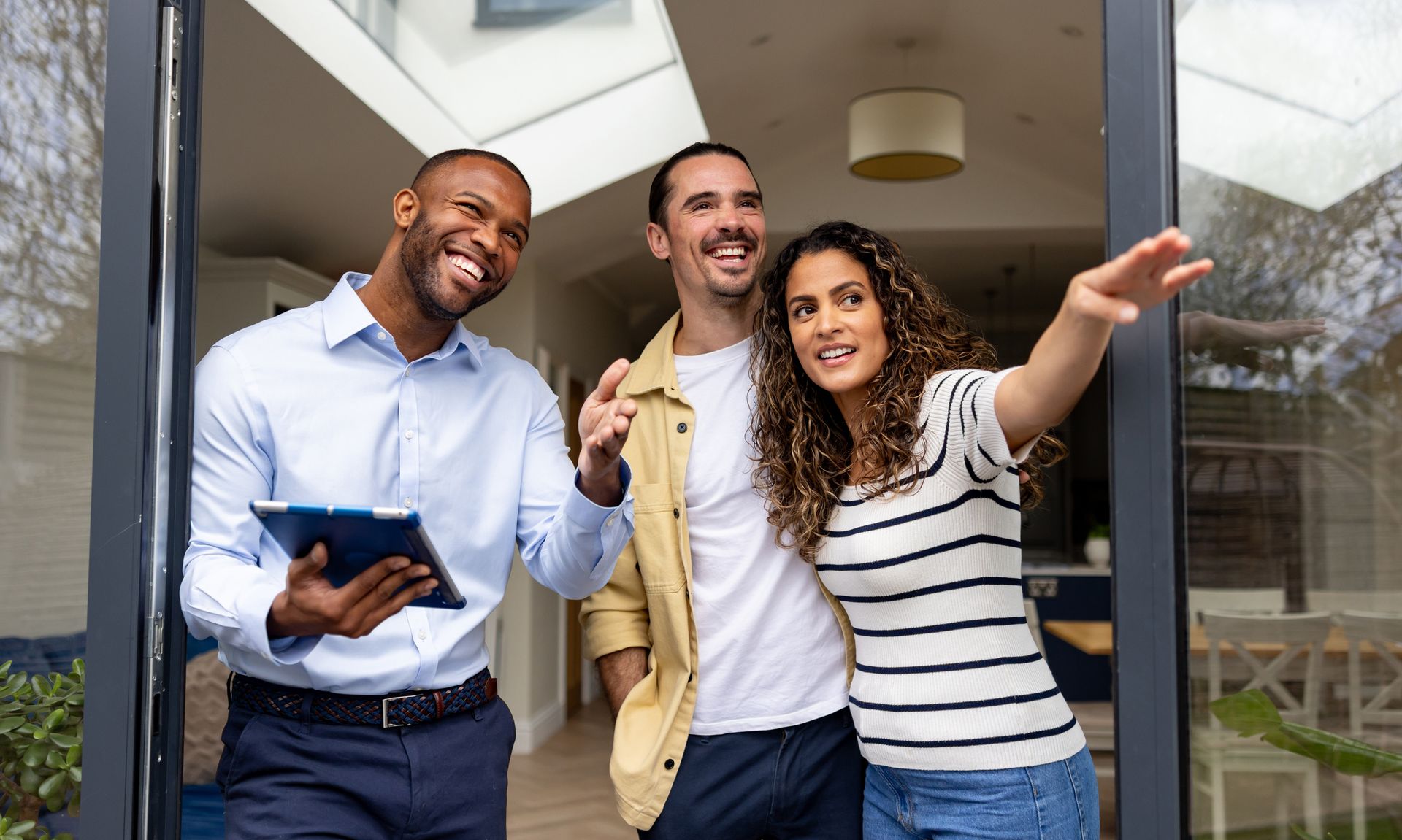 Real estate agent with couple viewing property; pointing towards something outside, smiling.