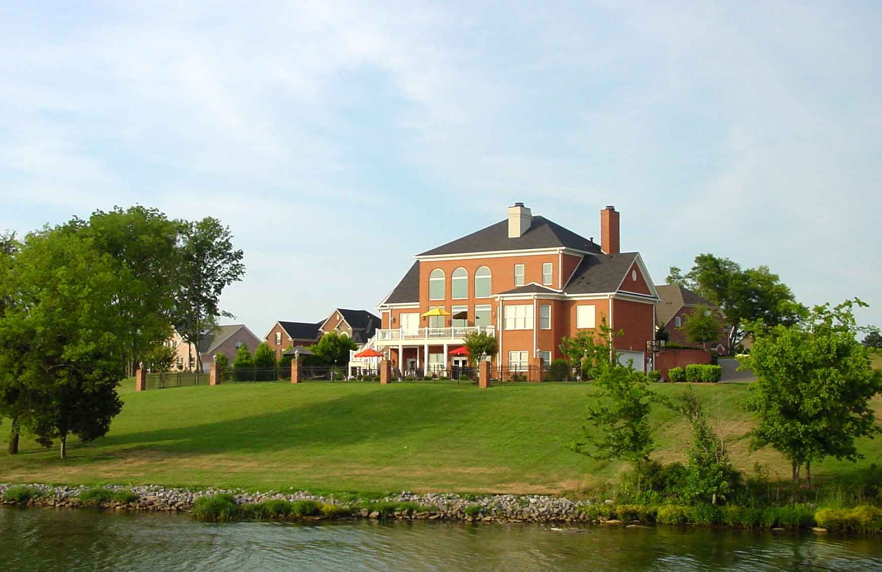 Large brick house with a white porch overlooking a grassy lawn and lake on a sunny day.