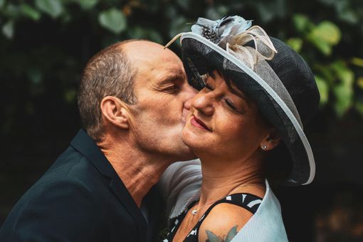 Man kissing a woman's cheek. She wears a hat with feathers, he wears a dark jacket. Both smile. Blurred outdoor background.