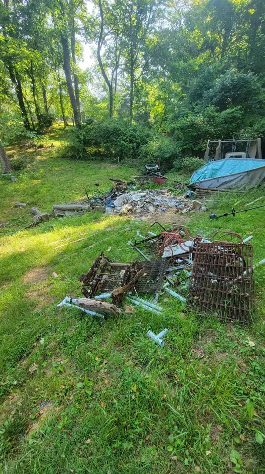 Grassy backyard with scattered debris. In the background, trees and a tarp-covered structure; foreground features metal and broken items.