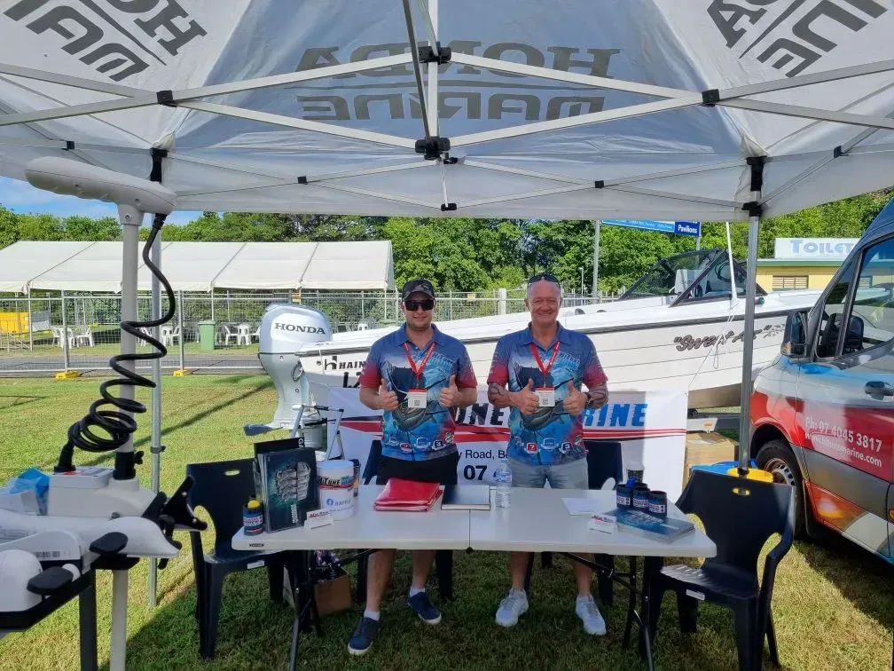 Two Men at A Honda Marine Booth, Holding Brochures, with A Boat in The Background — HiTune Marine In Port Douglas, QLD 