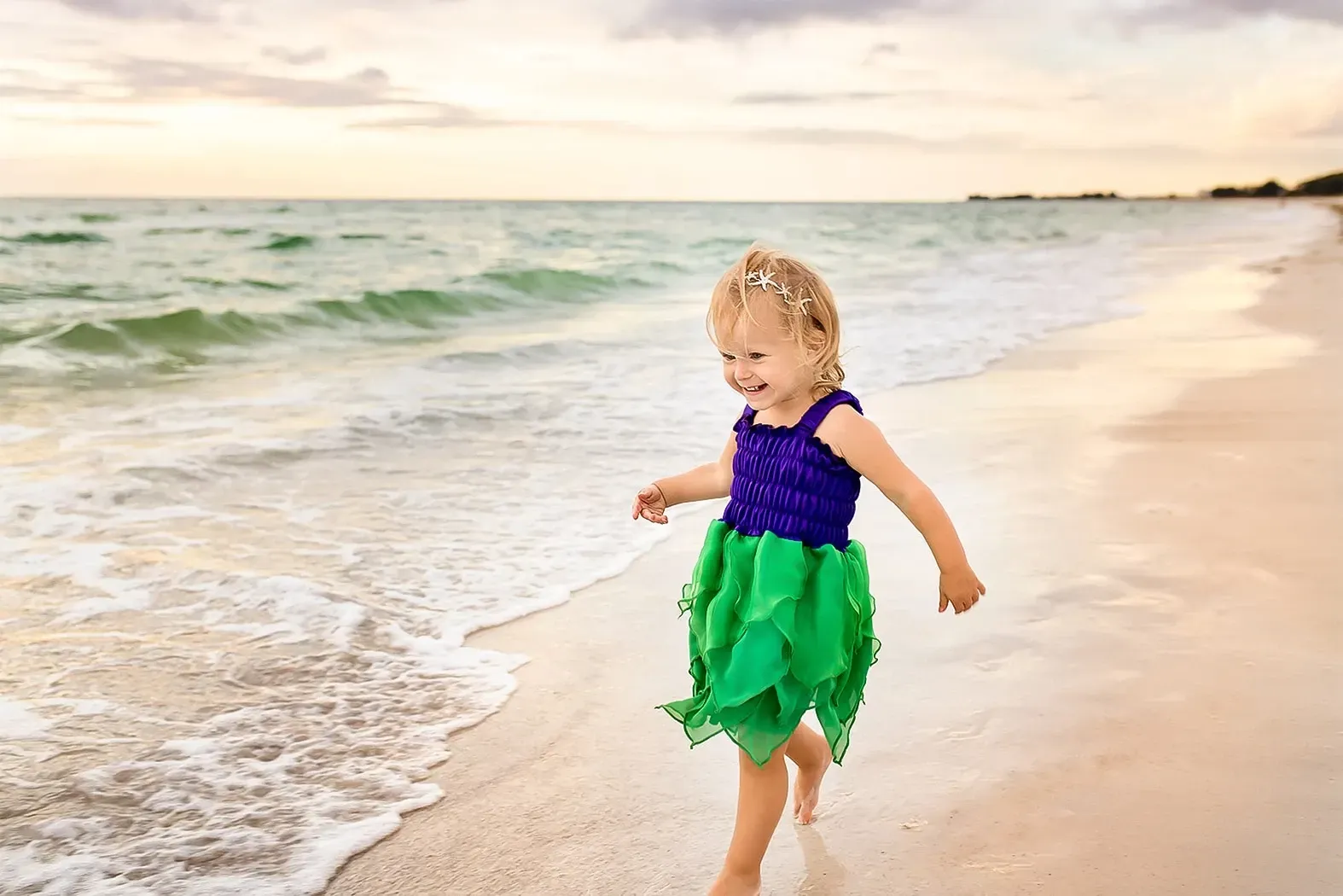 A little girl in a green and purple dress is running on the beach.