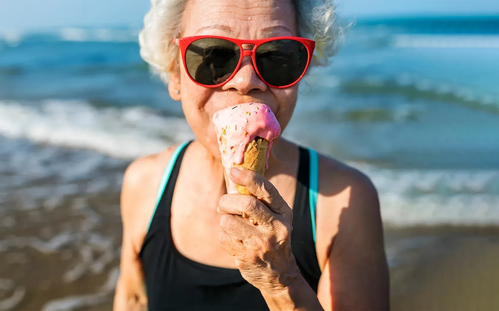 An elderly woman wearing sunglasses is eating an ice cream cone on the beach.