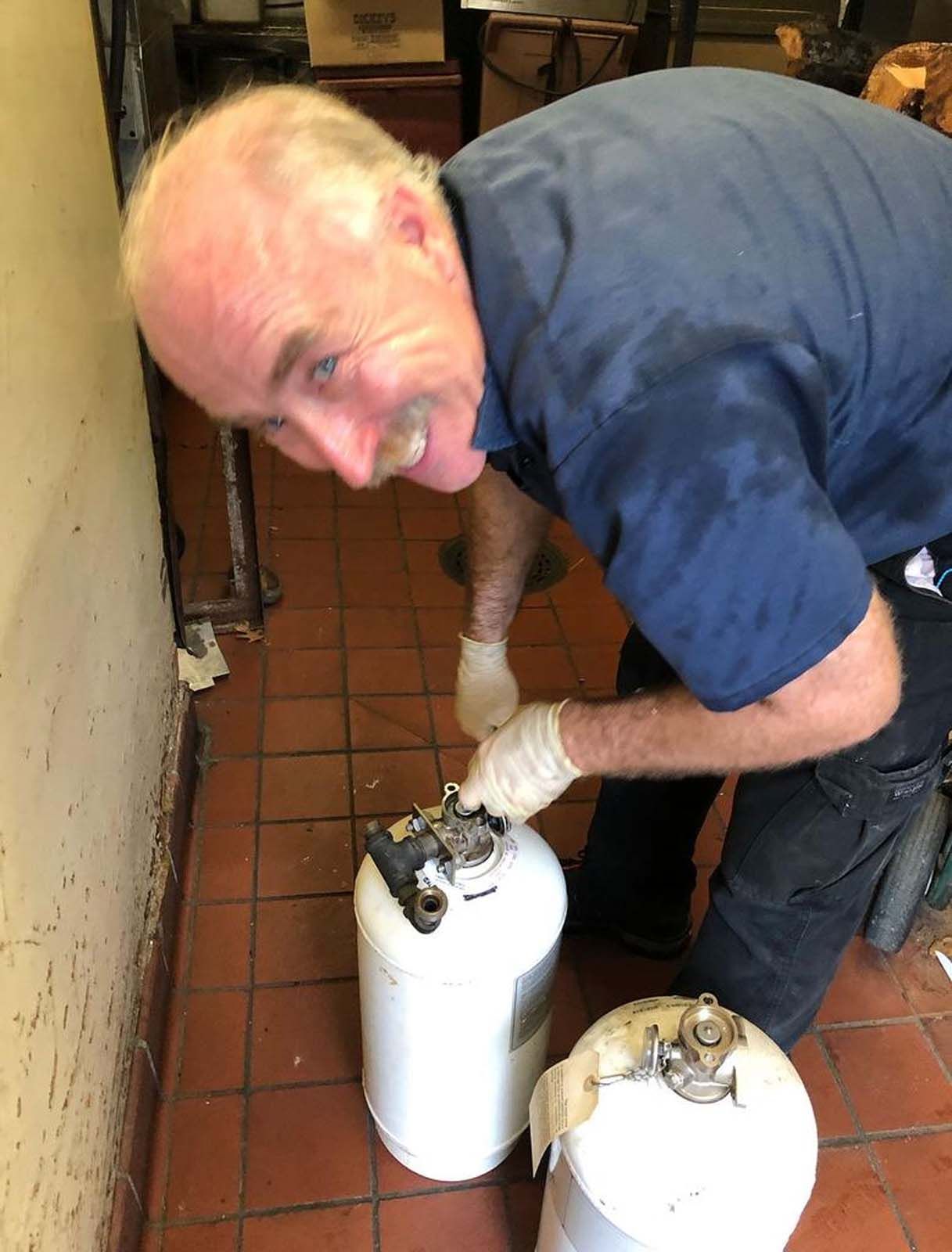 Man in blue shirt and gloves adjusts propane tank in a room with tiled floor.