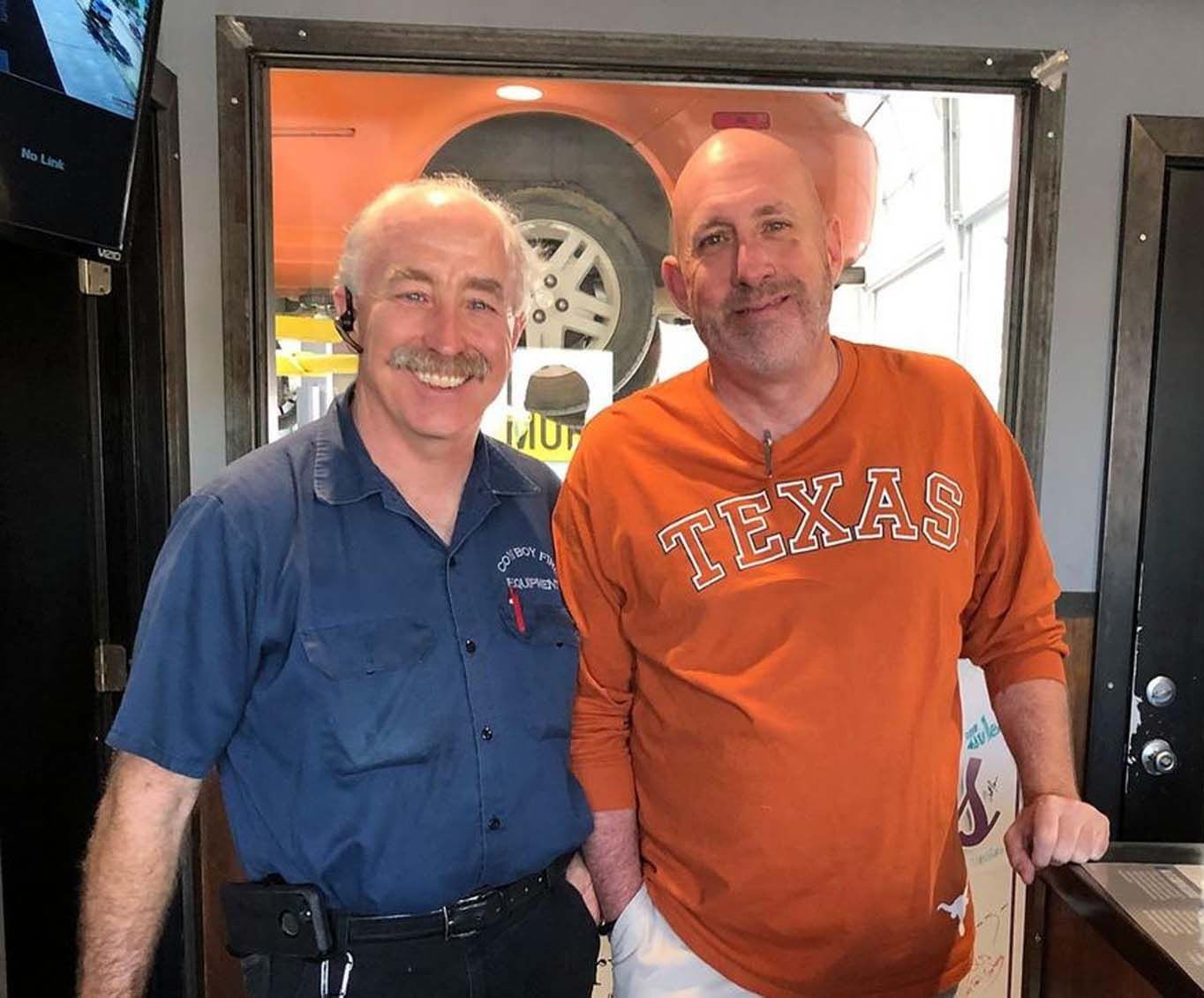 Two men smiling in a car repair shop. One wears a blue uniform, the other a burnt orange Texas Longhorns shirt.
