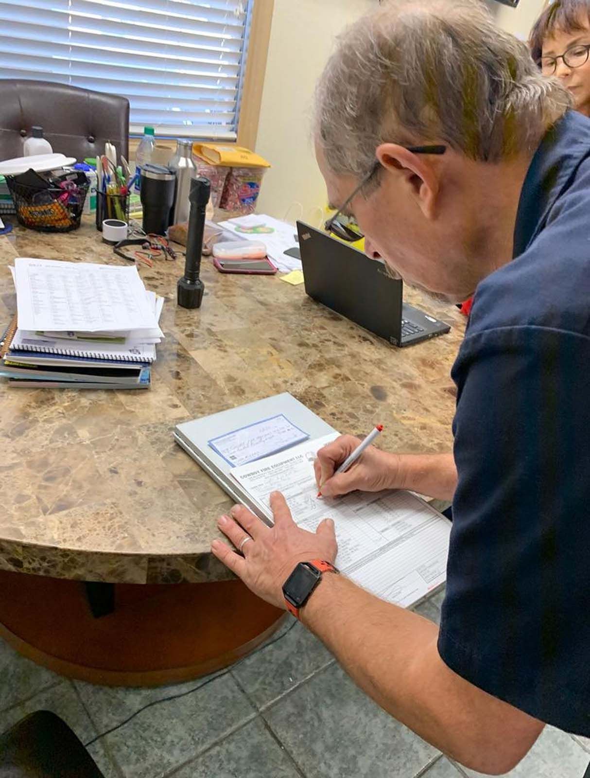 Person in blue shirt writing on a large document at a desk with a laptop and papers. Another person looks on.