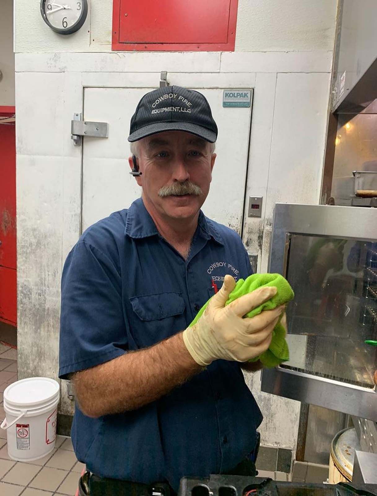 Man in a kitchen wearing a hat and gloves, holding a green cloth, near a stainless steel appliance.