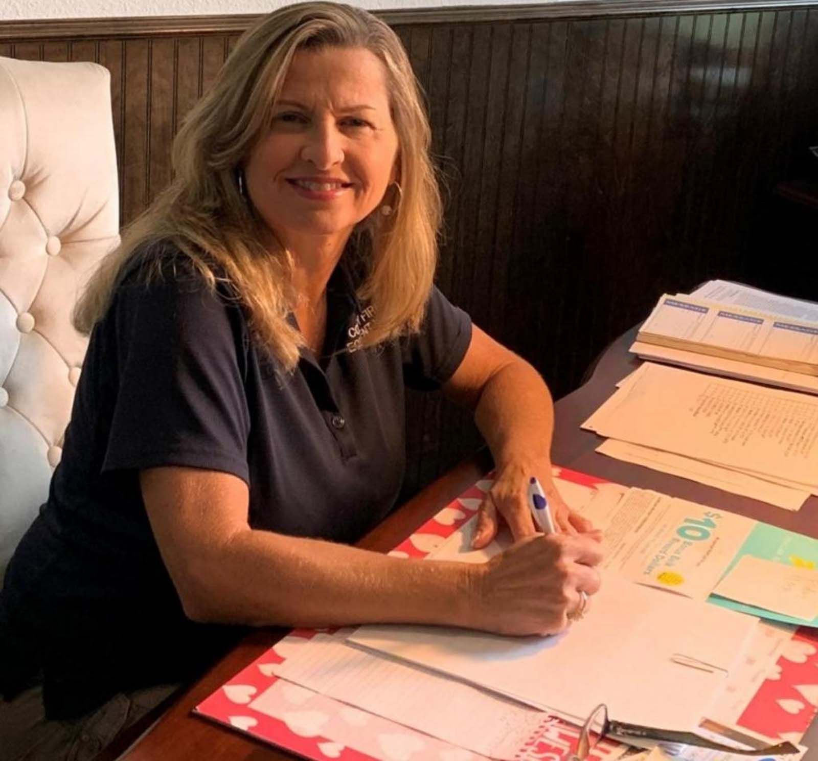 Woman with blonde hair signing documents at a desk, smiling. Dark shirt, light-colored chair.