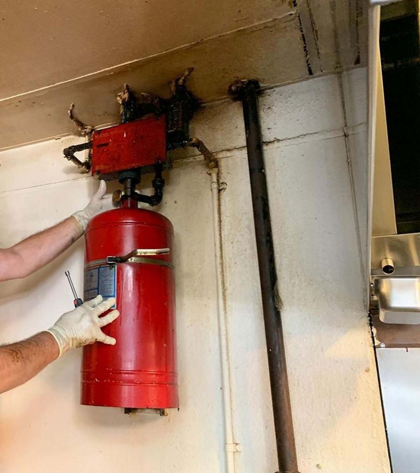 Hands wearing gloves checking a red fire suppression system in a commercial kitchen.