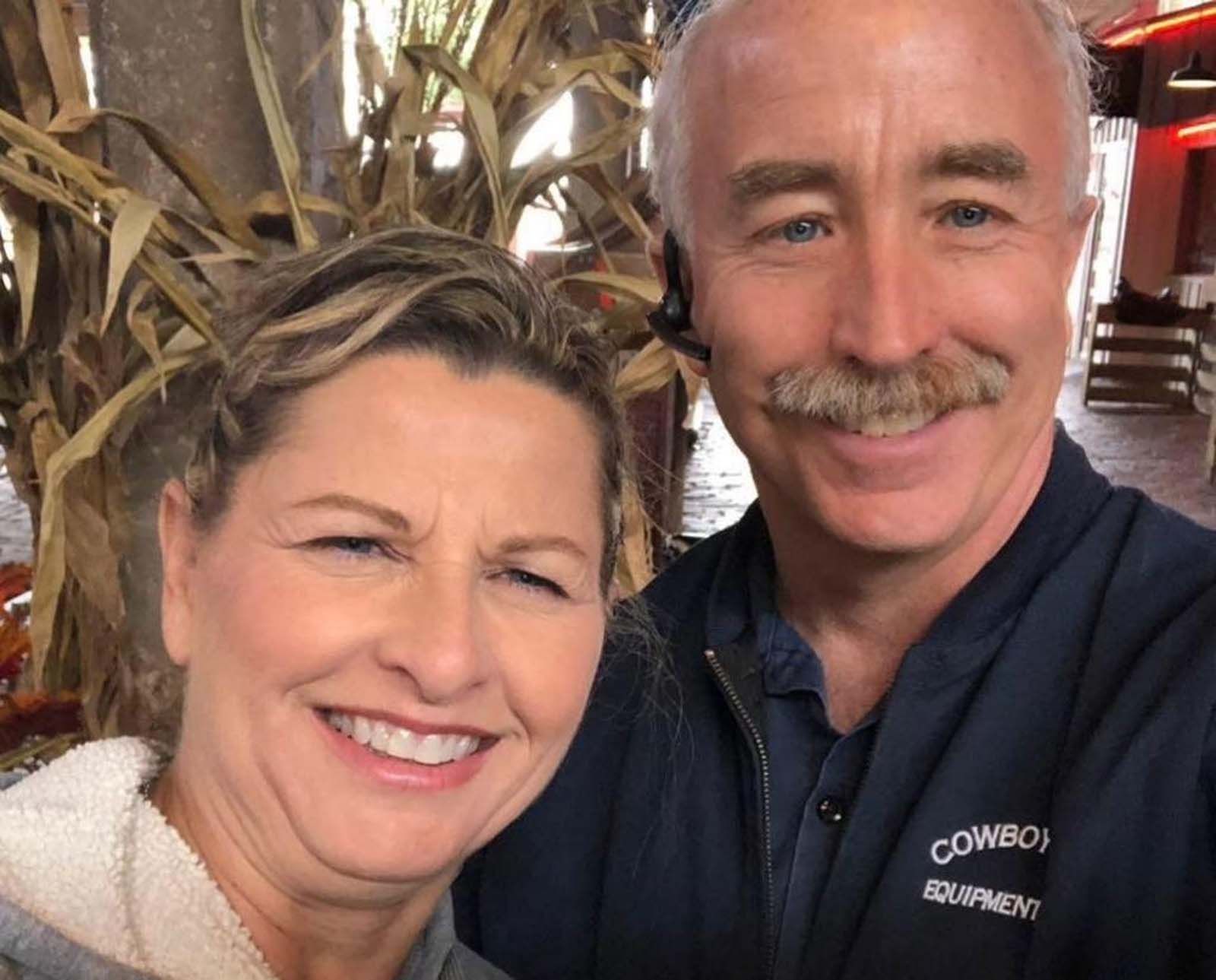 A woman and a man smile for a selfie in front of a rustic background with corn stalks.