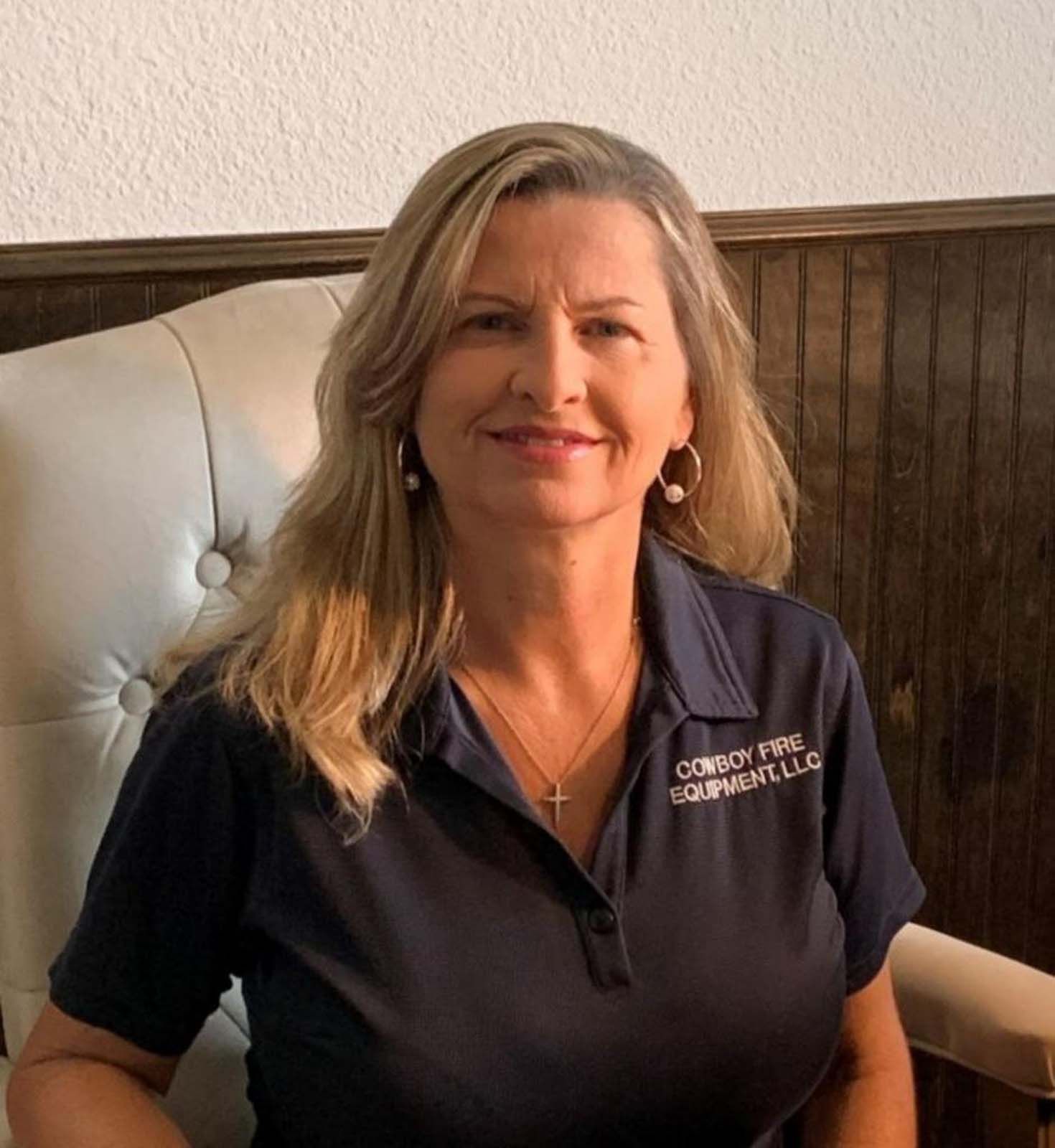 Woman in a navy polo shirt sits in a white chair. She has blonde hair and a cross necklace.