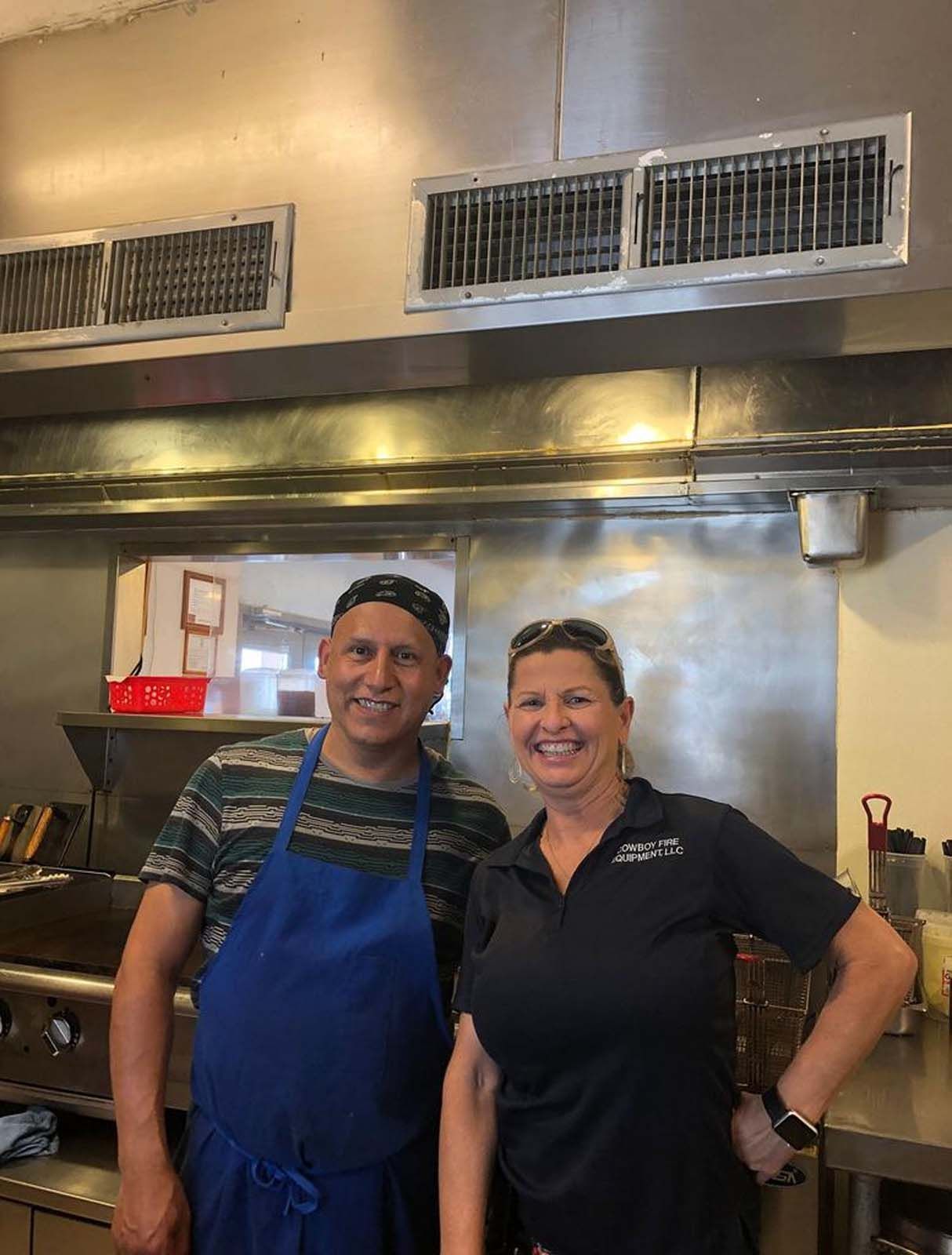 Two people in a commercial kitchen: a man in a blue apron and a woman in a black shirt, smiling
