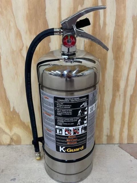 Stainless steel K-Guard fire extinguisher with black hose against a wood-grain background.