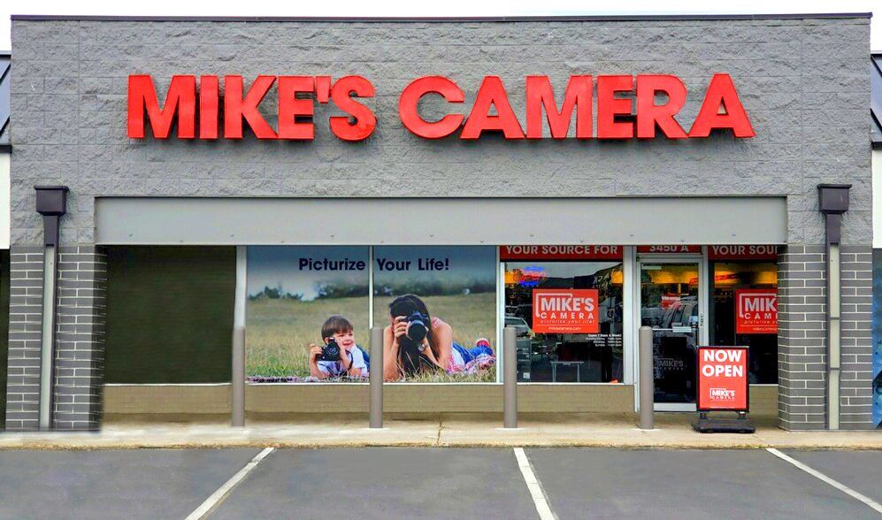 Mike's Camera storefront with red lettering, advertising photos. A couple is depicted taking pictures in an outdoor setting.