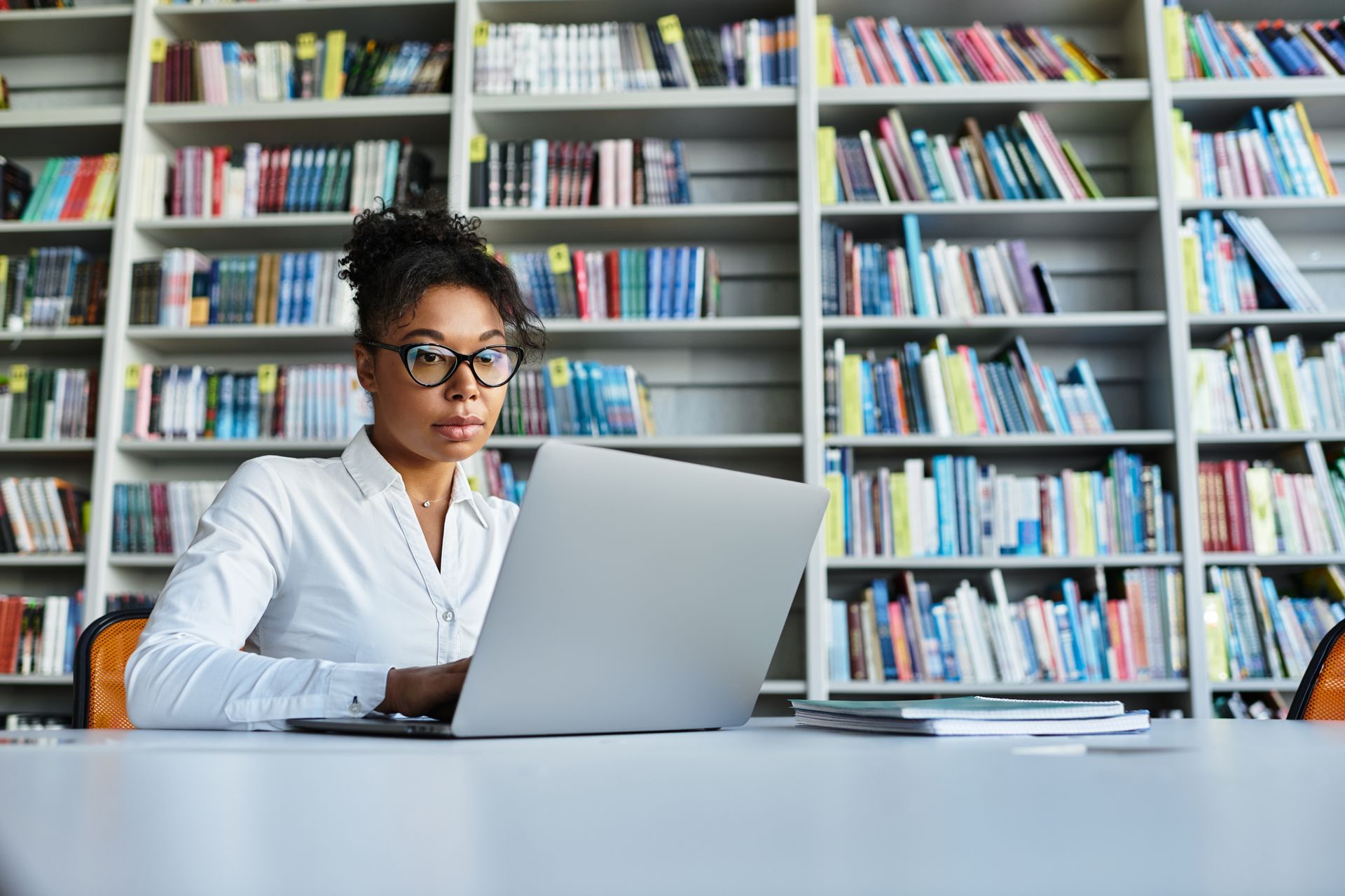 A female presenting person is sitting at a desk in a library using a laptop computer.