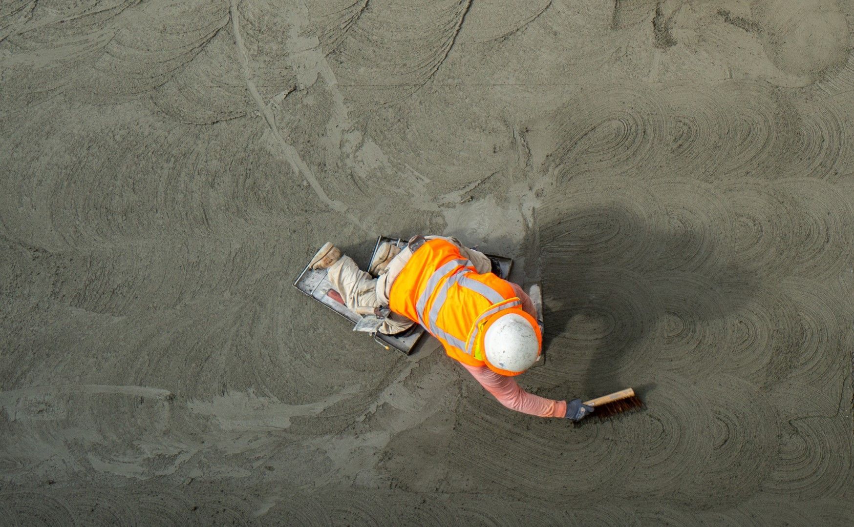 Construction worker smoothing concrete, wearing orange vest and white helmet.