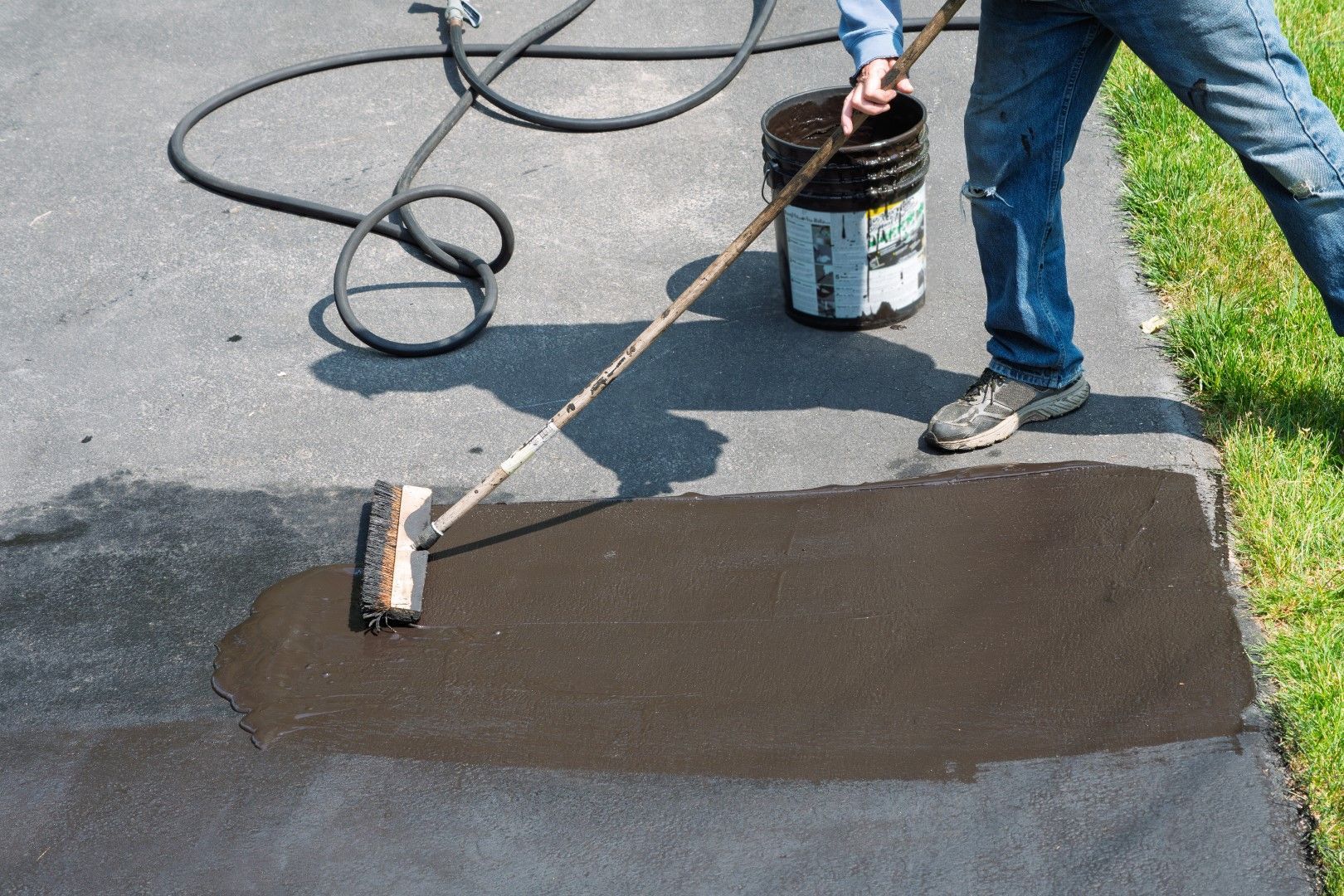 Person applying black asphalt sealant to a driveway with a roller. A bucket of sealant is nearby.
