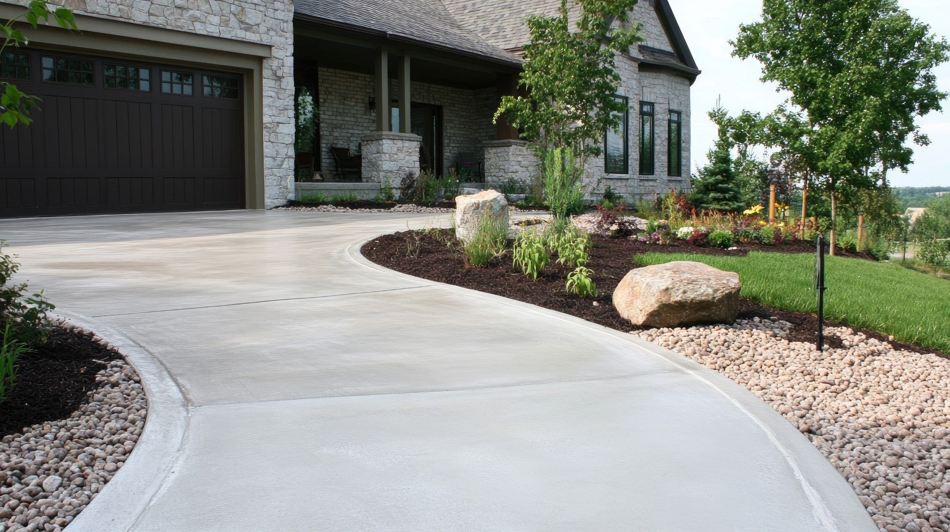 Curving concrete driveway leading to a modern house with landscaped yard, brown garage door, and trees.