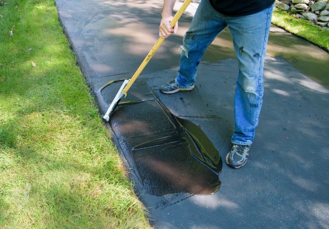 Person applying black sealant to an asphalt driveway with a long-handled squeegee.