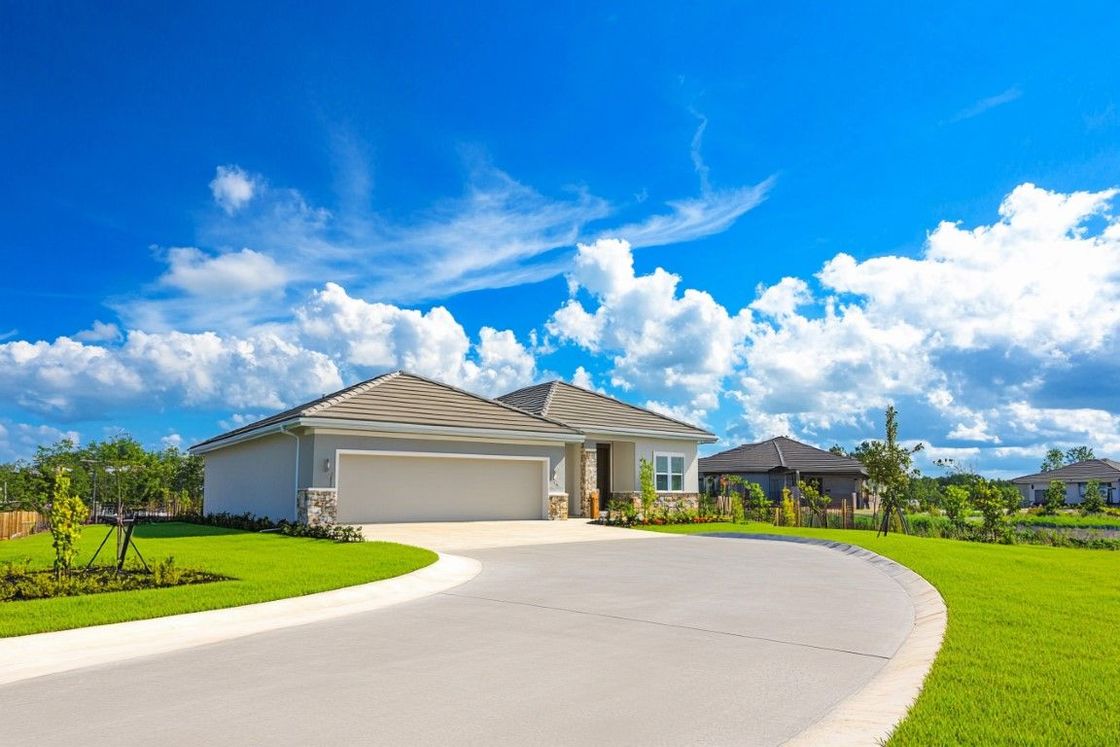 House with a curved driveway, surrounded by green lawn under a bright blue sky with fluffy white clouds.