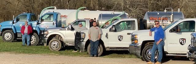 Four people standing in front of four work trucks, including two vacuum tank trucks, parked on a grassy area.