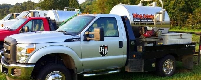 A silver utility truck with a mounted metal tank and KJ logo, parked outdoors with a red vehicle visible in the background.