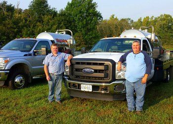 Two people stand in a field in front of two white service trucks with large tanks mounted in their beds.