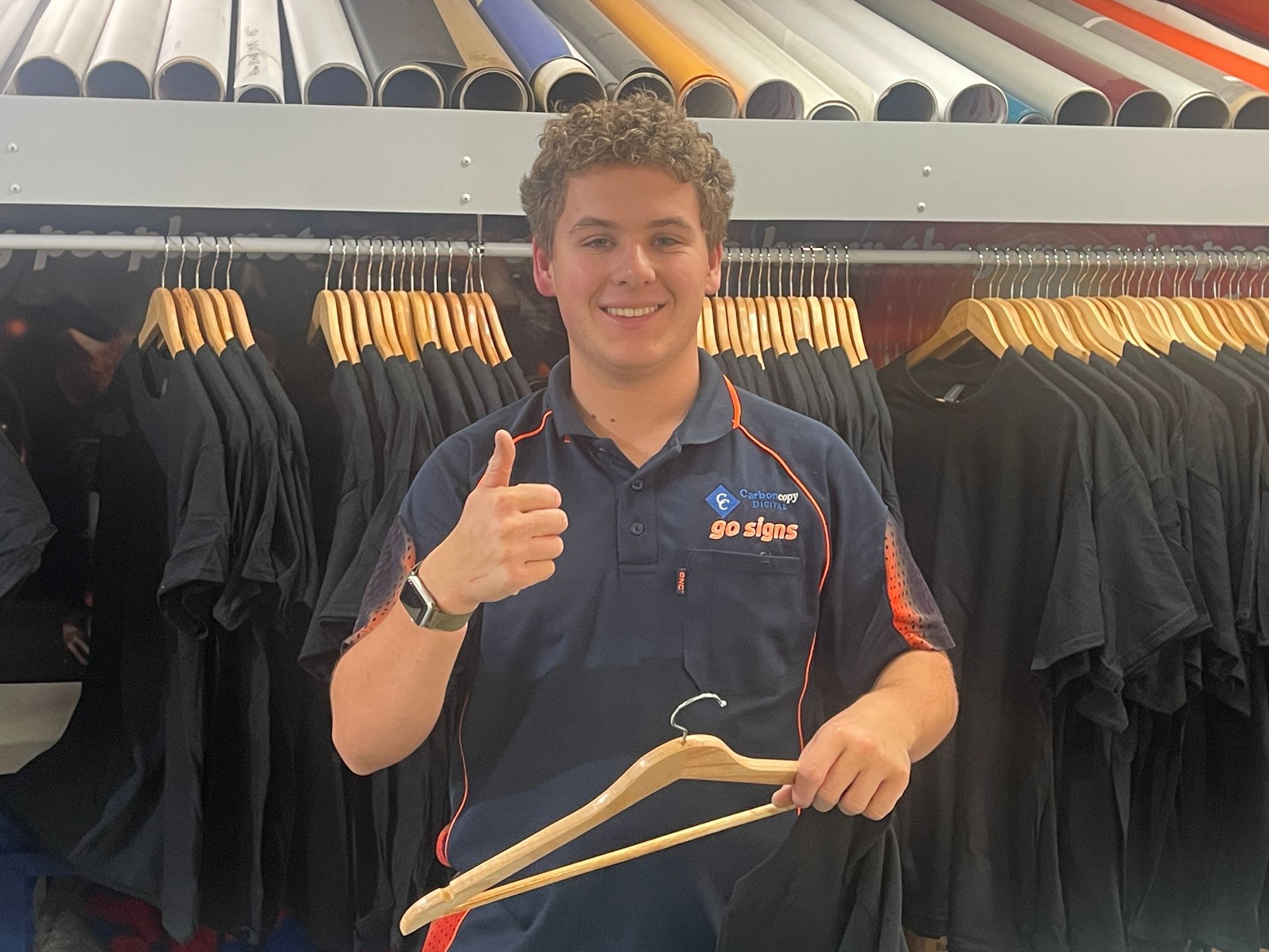 Person in blue polo gives thumbs-up, holding a wooden hanger in a clothing store; racks of black shirts in the background — Carbon Copy in Port Macquarie, NSW