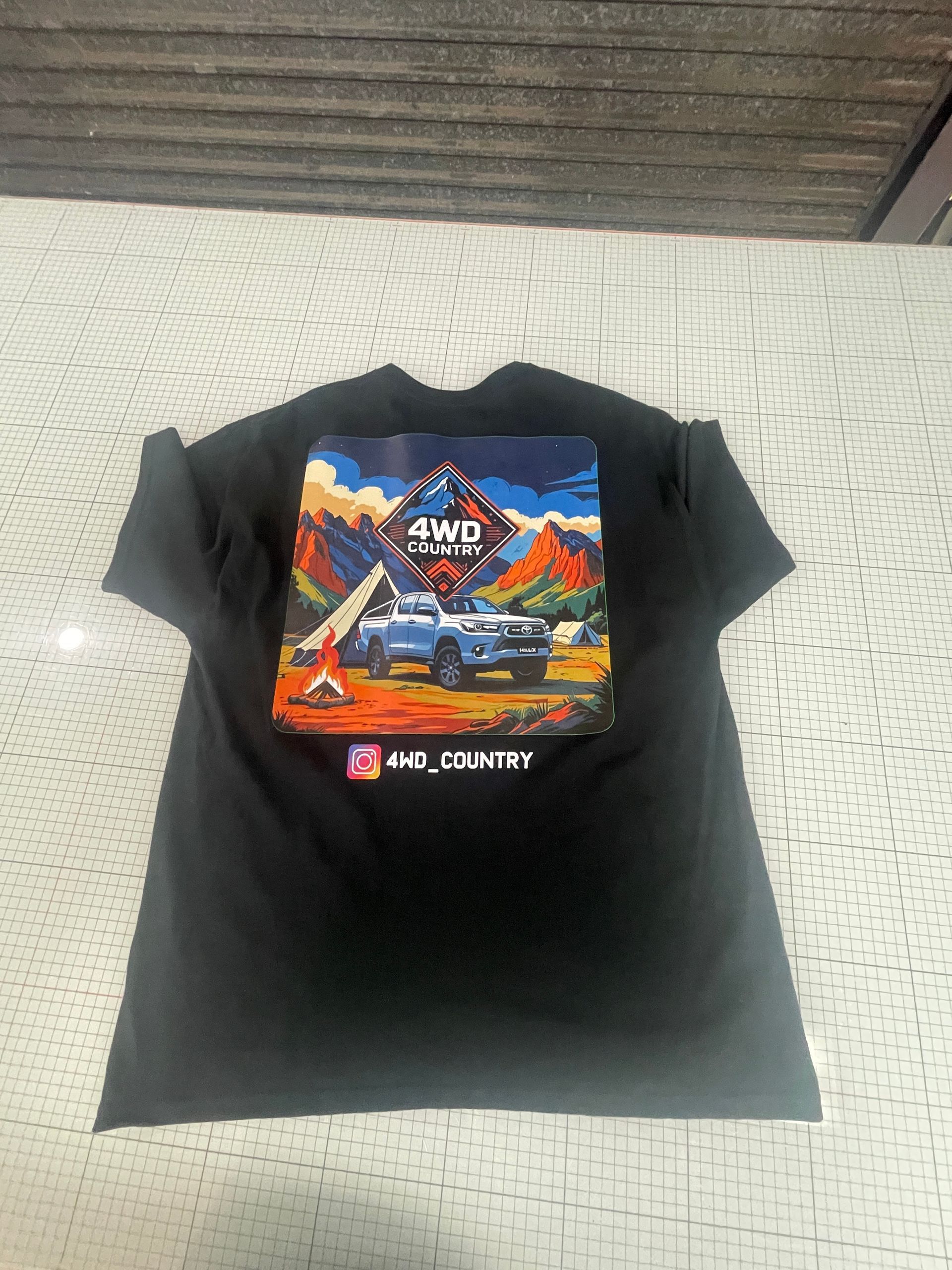 Black t-shirt with a blue 4WD truck graphic on the back. The background shows mountains and desert landscape — Carbon Copy in Port Macquarie, NSW