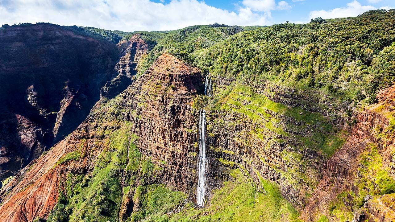 Manawaiopuna Falls, also known as Jurassic Falls, cascading 400 feet down a lush cliffside in Hanapēpē Valley on Kauai, viewed from a scenic helicopter tour.