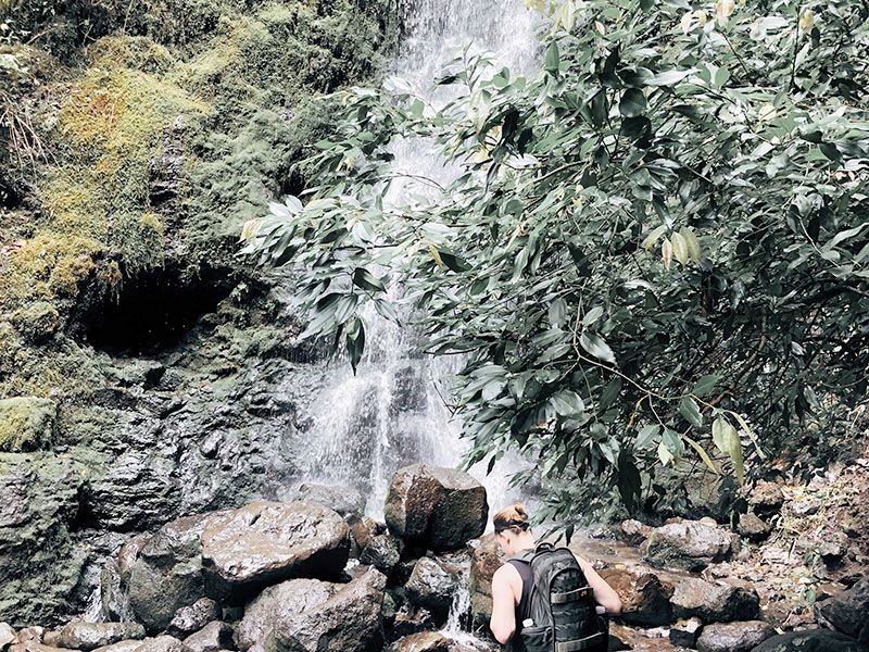 A woman is standing next to a waterfall in the woods.