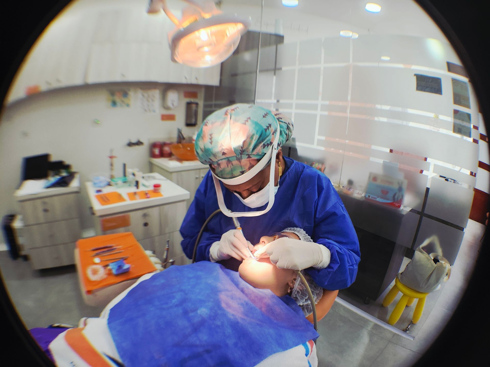 Dentista con bata azul y mascarilla atendiendo a un paciente en una clínica dental, con instrumental quirúrgico y luces brillantes en el techo.