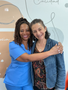 Dos mujeres sonriendo y abrazándose frente a una pared azul claro con escritura cursiva.