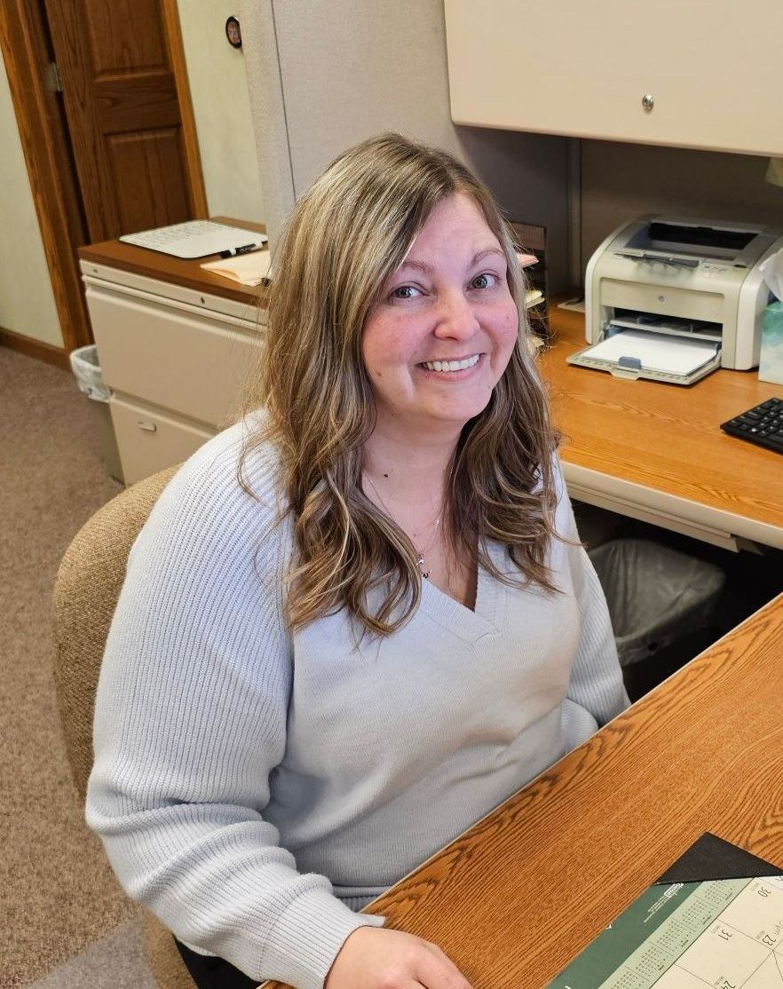 A woman is smiling while sitting at a desk in front of a printer.