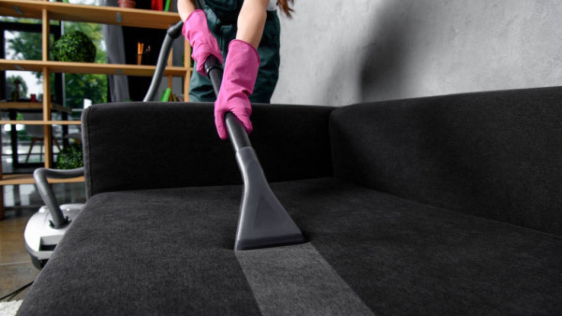 A woman is cleaning a black couch with a vacuum cleaner.