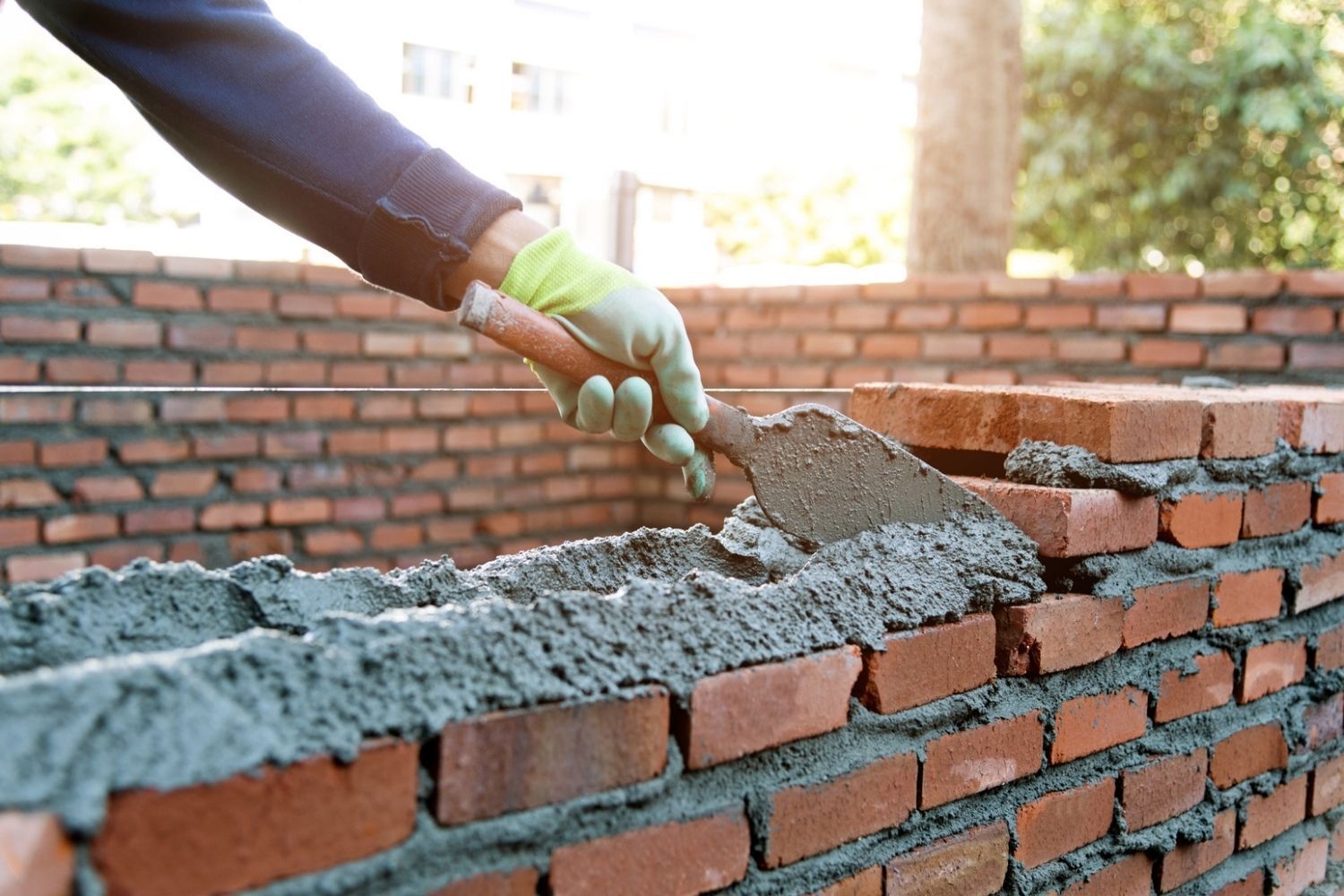 A gloved hand uses a trowel to apply mortar between red bricks, building a wall outdoors.