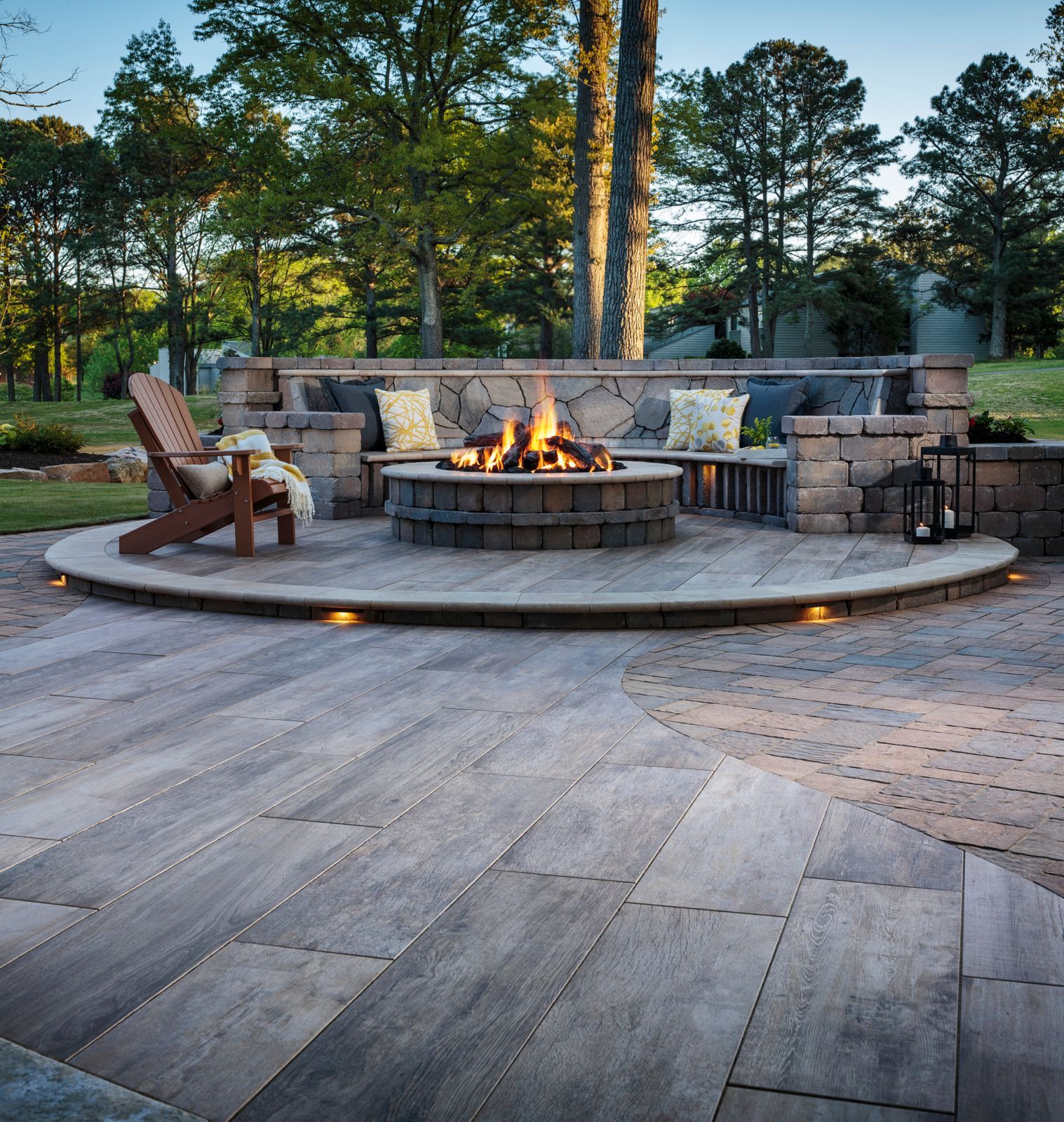 Outdoor fire pit with seating on a stone patio, surrounded by trees and a green lawn.