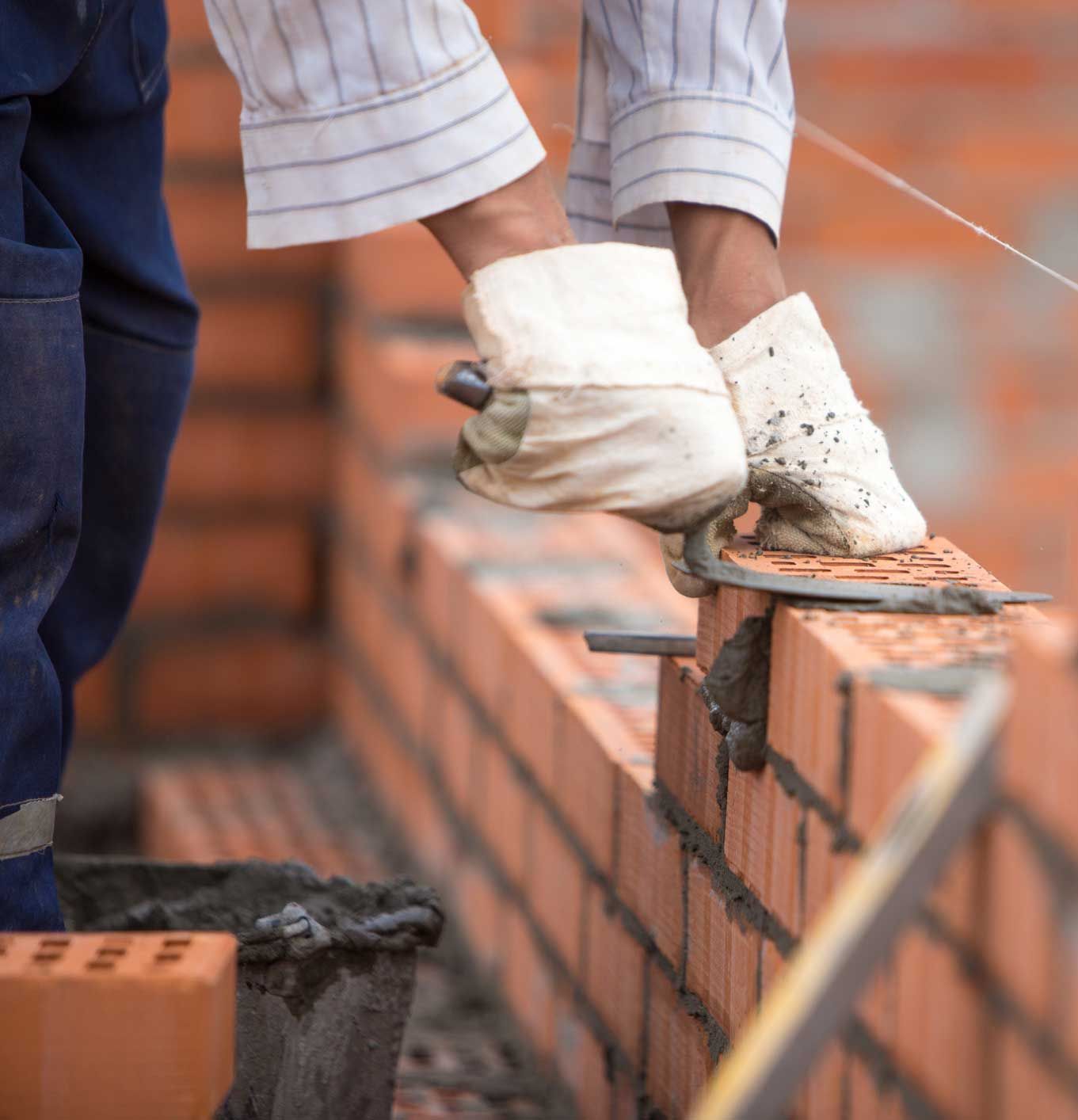 A person wearing gloves using a trowel to lay bricks on a partially built brick wall.