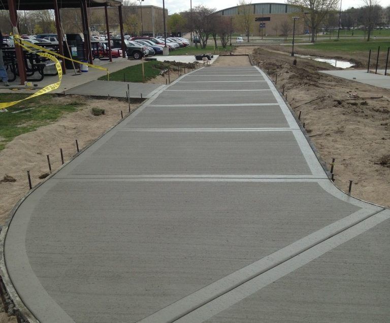 Newly poured concrete sidewalk with expansion joints curving to the left. Construction area with parking lot in the background.