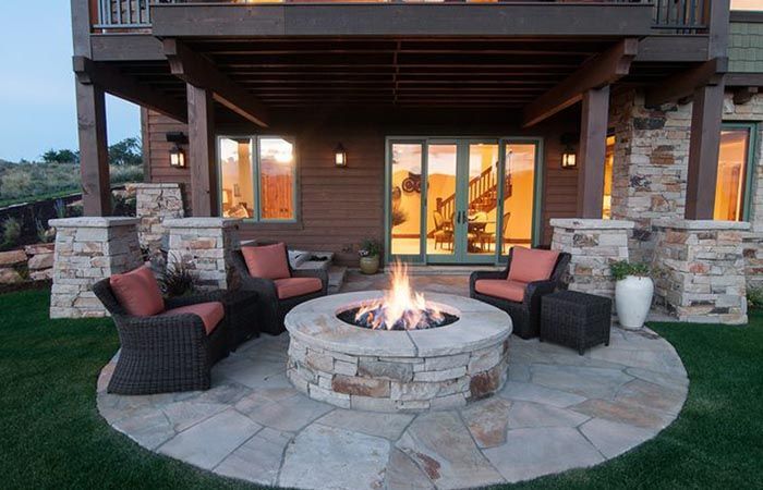 Stone fire pit with chairs on a patio under a house with green views.