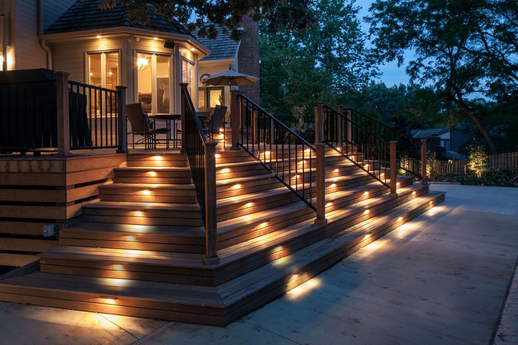 Outdoor wooden staircase lit by embedded lights, leading up to a deck and house at dusk.