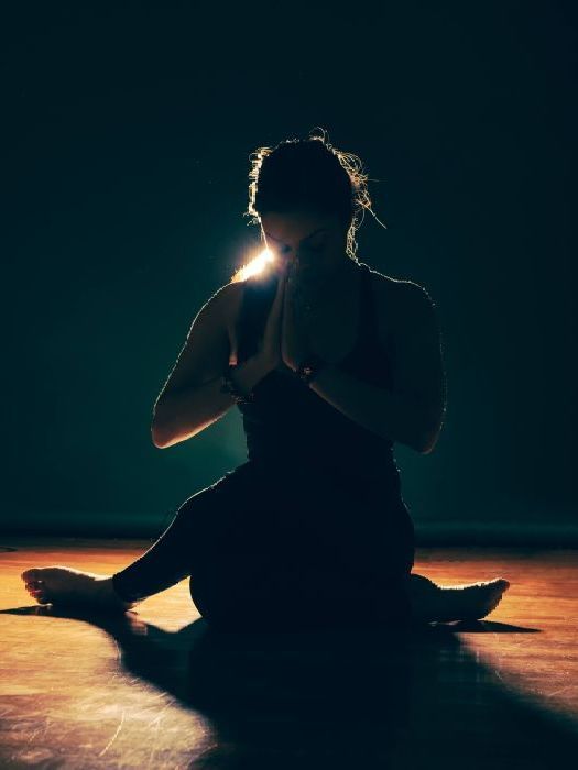 A woman is sitting on the floor in a dark room with her hands folded in prayer.