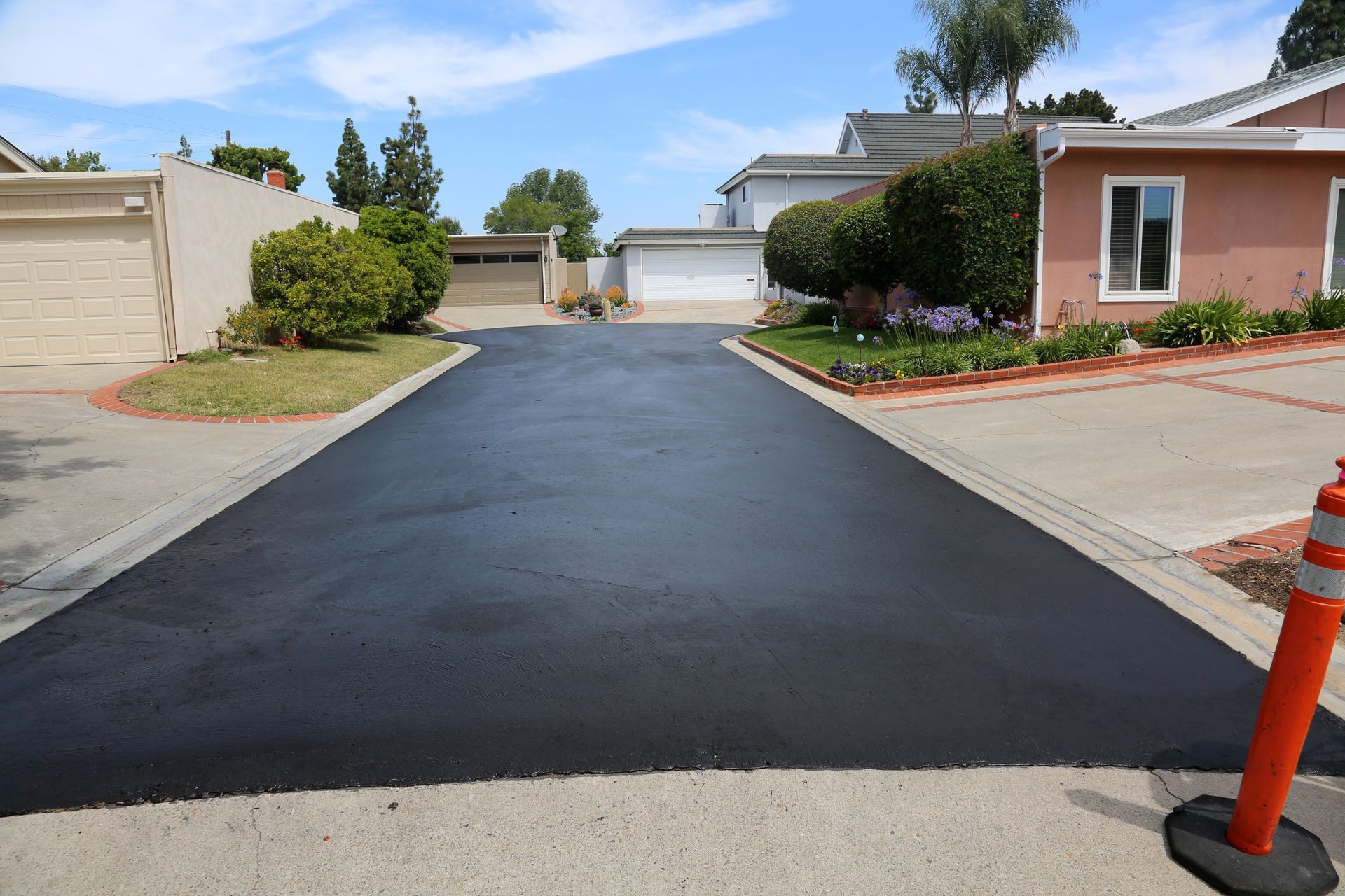 Paved driveway leading to houses with manicured lawns and blue sky. A traffic cone is on the right.