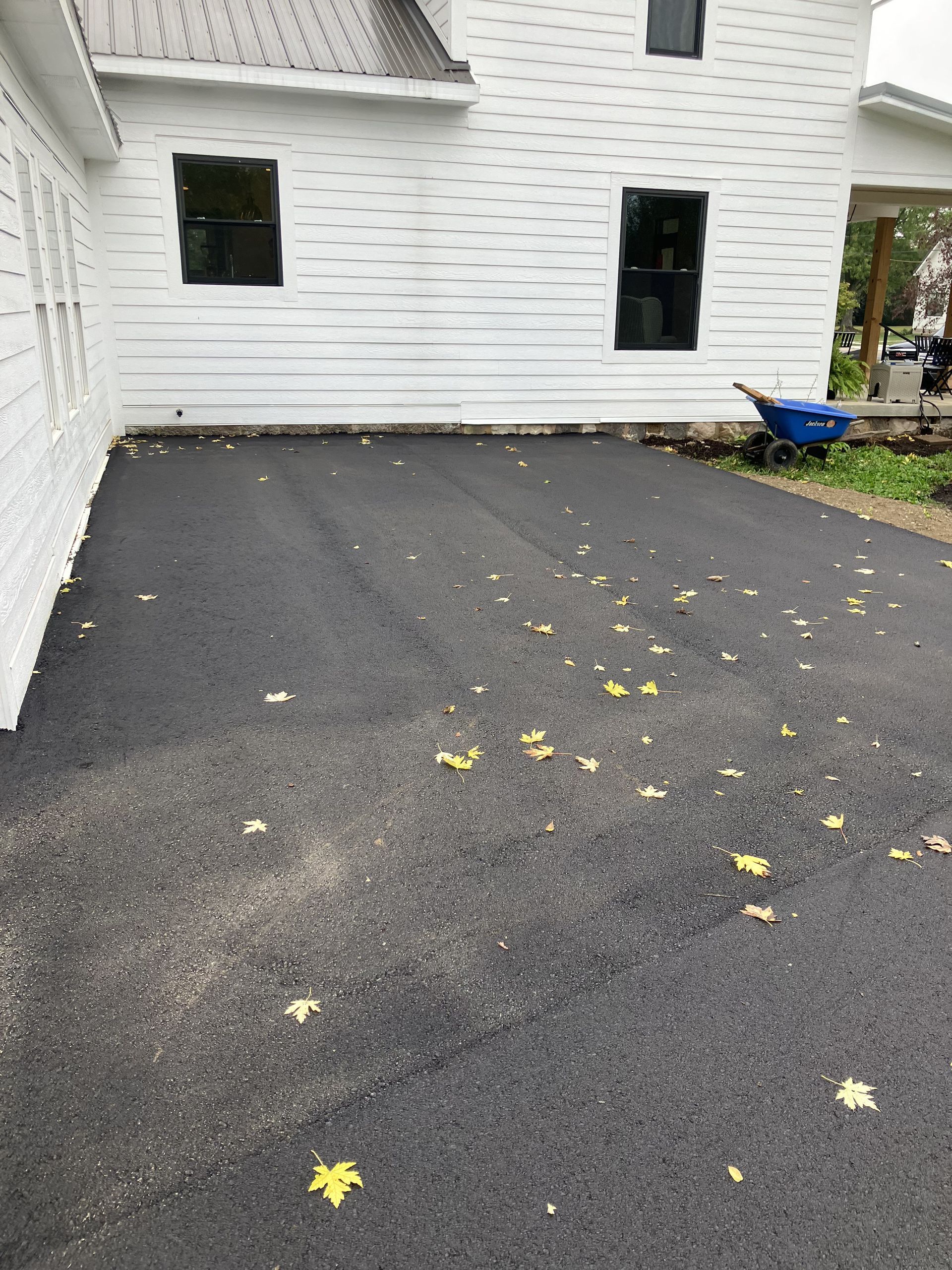 Newly paved blacktop driveway next to a white house with dark windows. Fallen yellow leaves scattered.