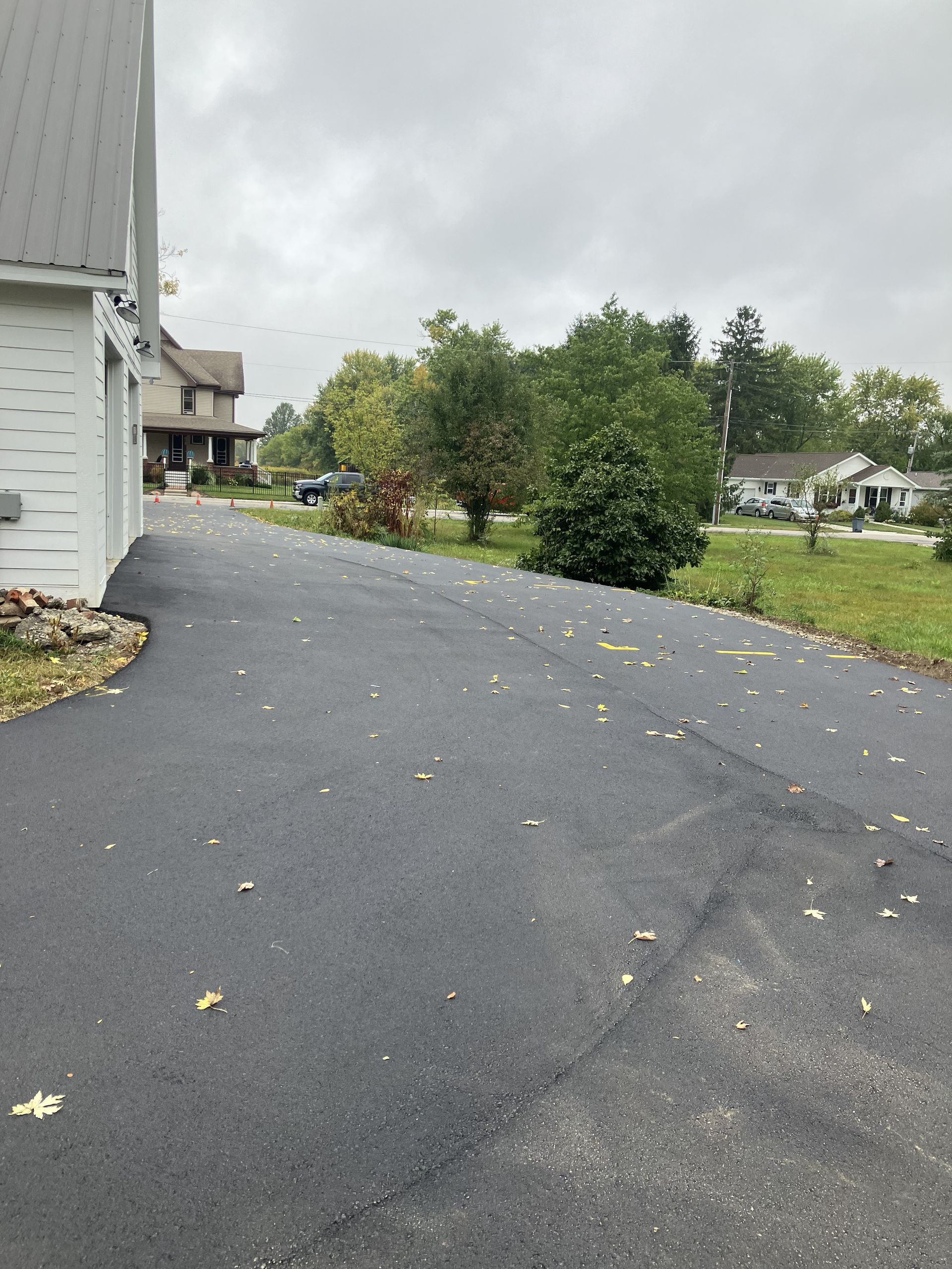 Black asphalt driveway with scattered leaves; houses and trees in the distance under a cloudy sky.