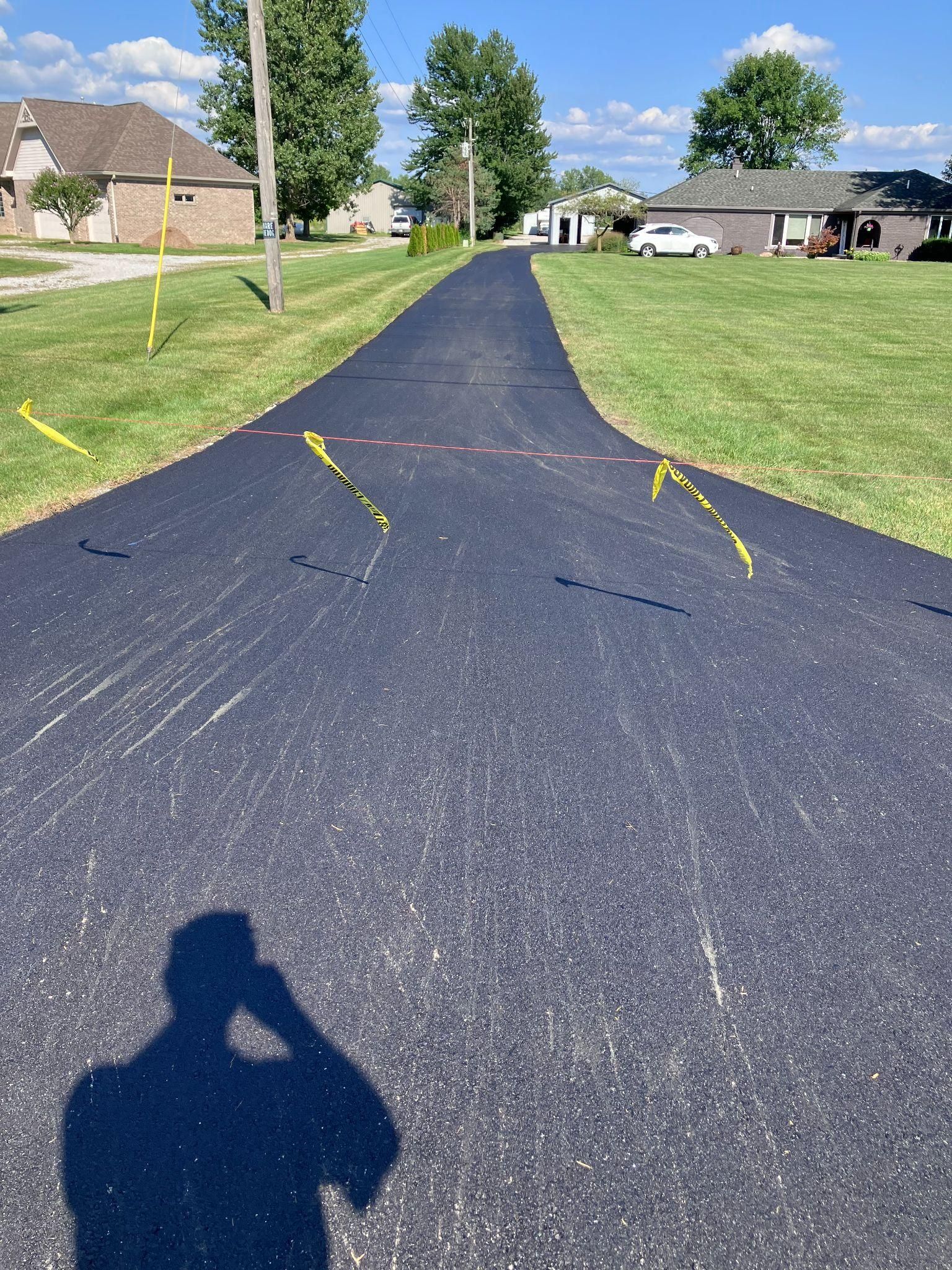 Newly paved asphalt driveway with yellow marking flags, green lawn, houses, and the shadow of a person.